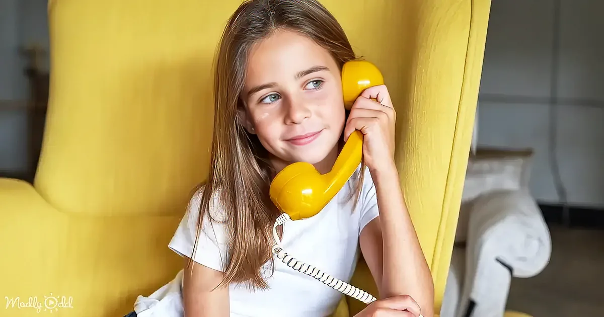 A girl with long brown hair sits on a yellow chair, holding a yellow telephone receiver to her ear.