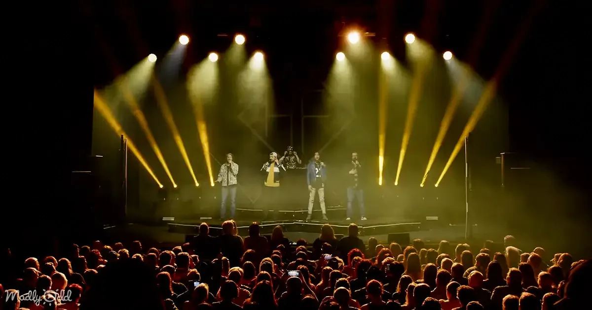 A group of performers on stage with bright yellow lights and a crowd in the foreground.