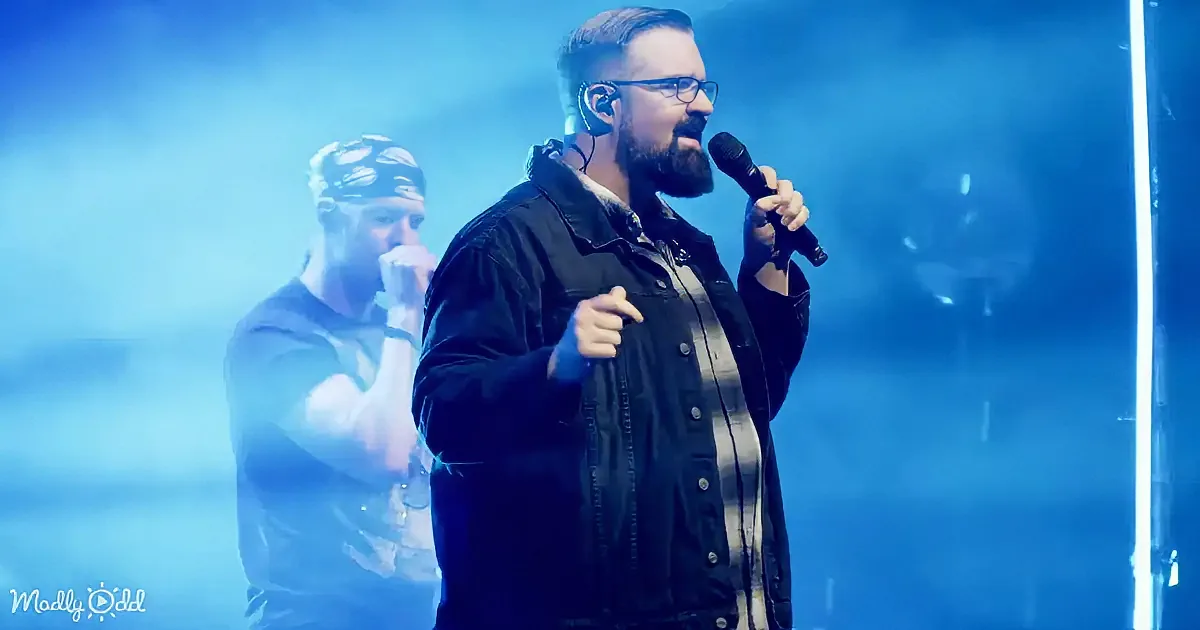 A man with a beard and glasses holds a microphone, gesturing on stage with a blue-lit background.