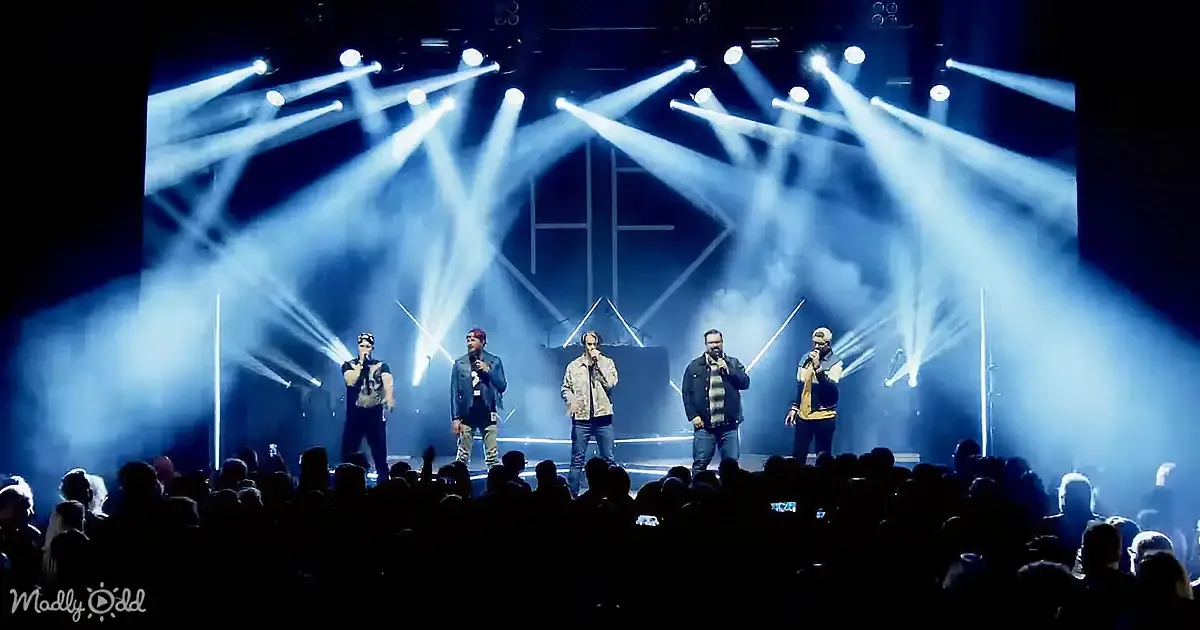 Six performers on stage with microphones, illuminated by bright stage lights and smoke, captivating an audience.