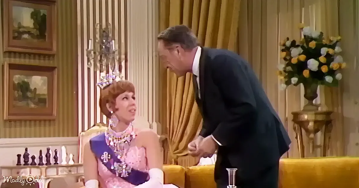 A man in a suit leans towards a woman in a pink dress with a sash and tiara, seated on a yellow couch.