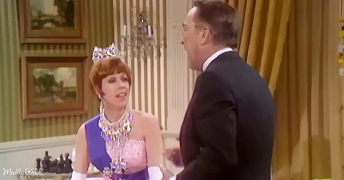 A woman in a purple dress and necklace is talking to a man in a suit in a room with striped wallpaper and paintings.