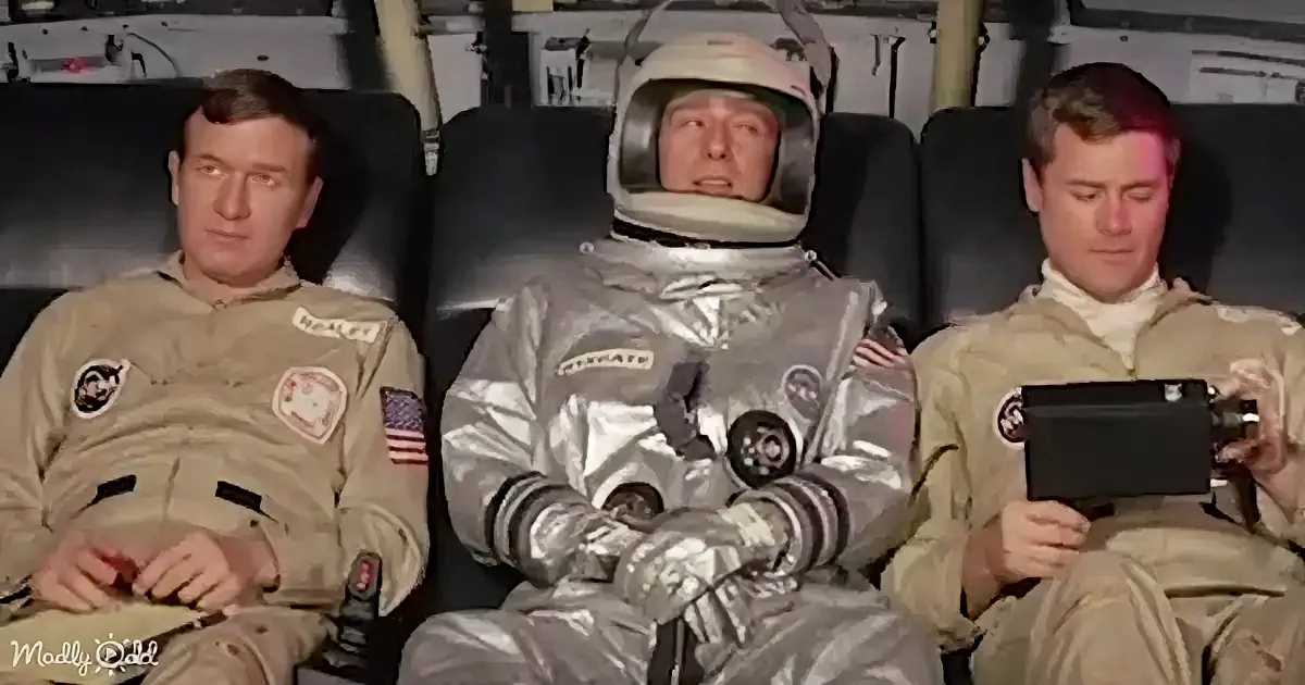 A man in a silver space suit sits between two men in tan uniforms, one holding a tablet.