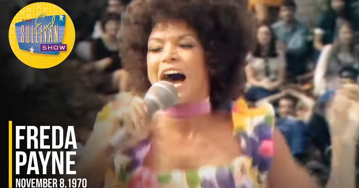 A smiling woman with a large afro holds a microphone, wearing a colorful outfit, with a band and audience in the background.