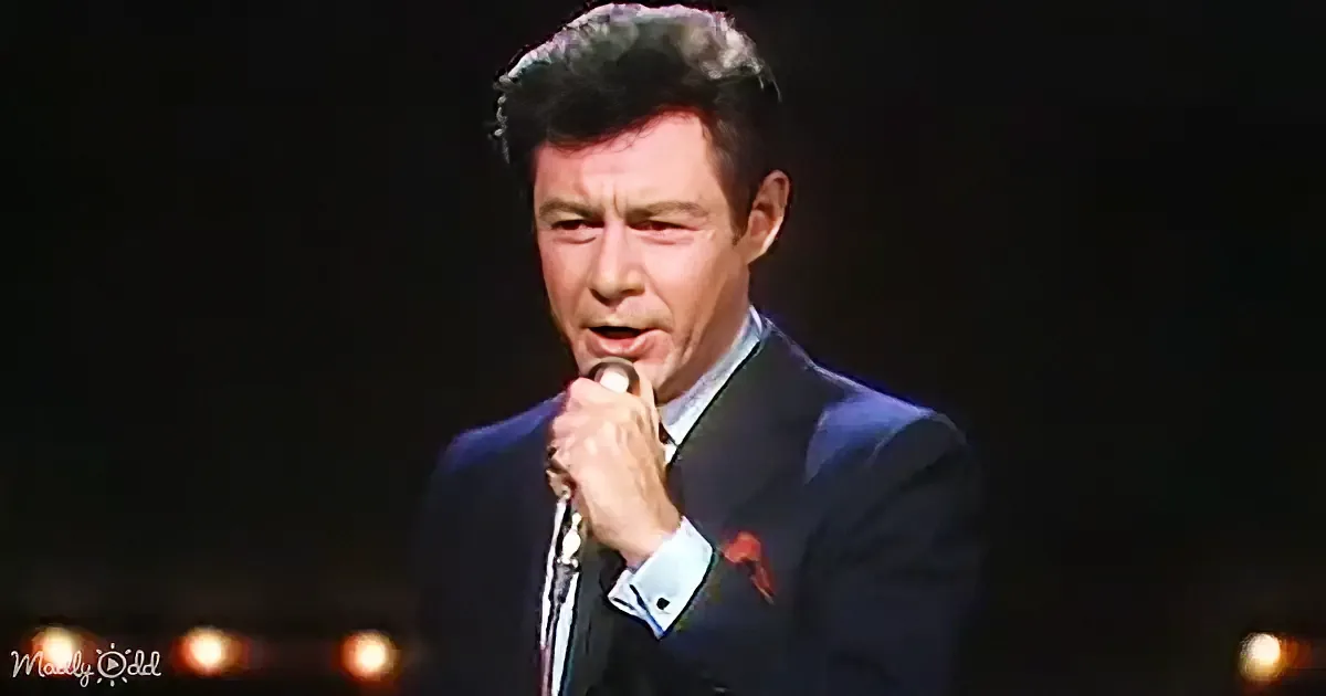 A man in a suit sings into a microphone on stage, with dim lighting and blurred background.