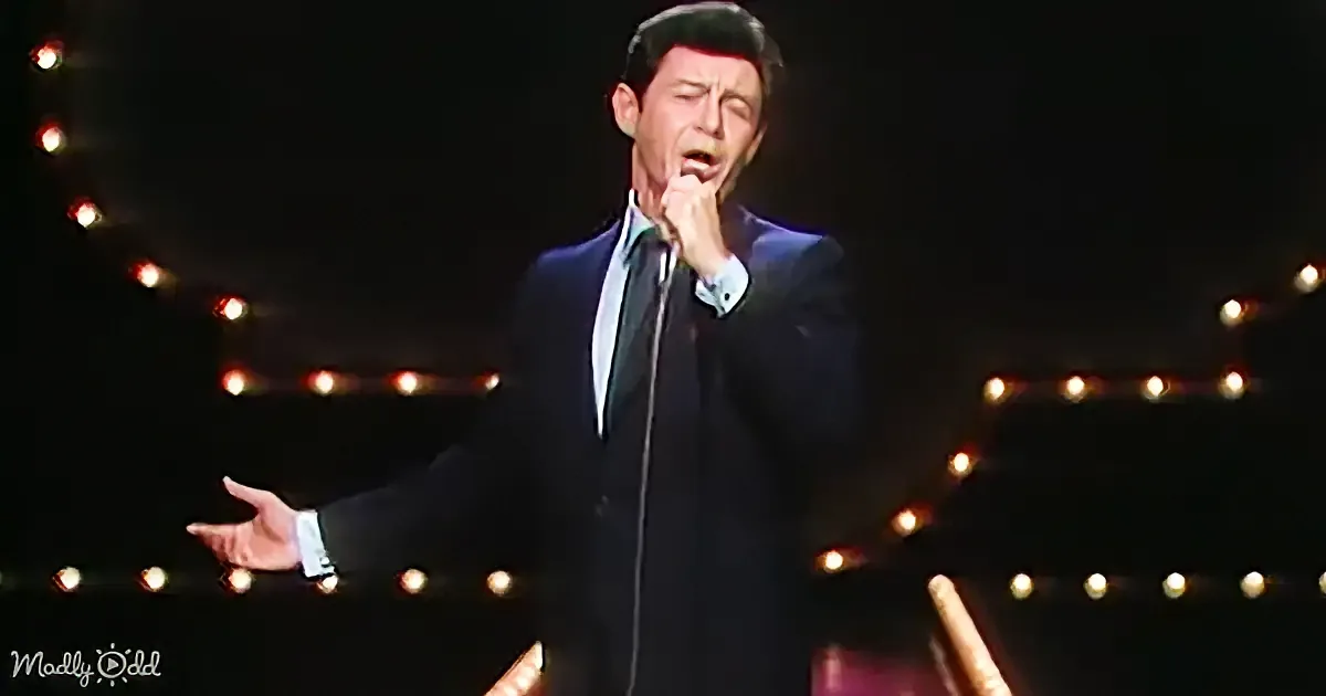 A man in a suit sings into a microphone on stage with string lights in the background.