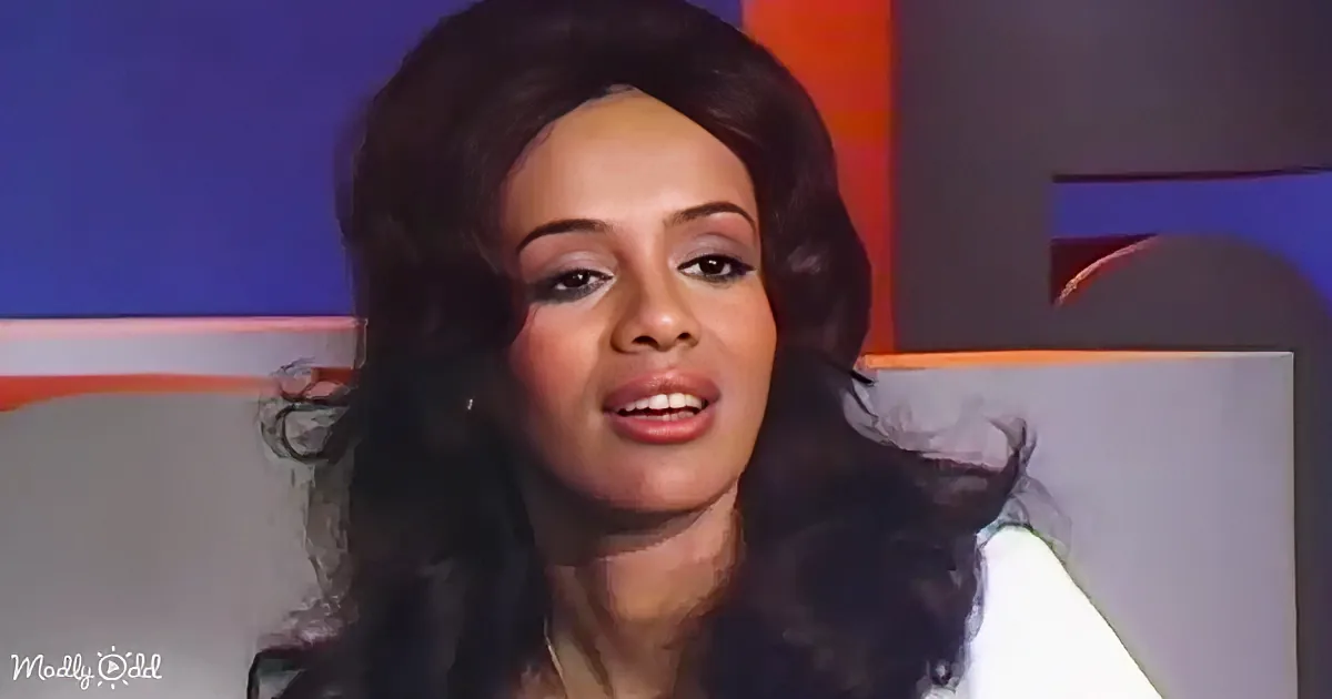 A woman with long, dark, curly hair is speaking in front of a colorful background.