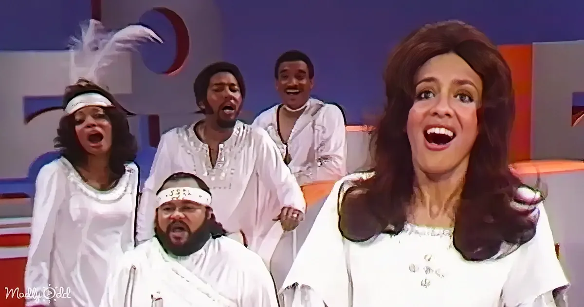 A woman in white attire sings passionately, surrounded by a group of men in similar costumes, all on a stage.