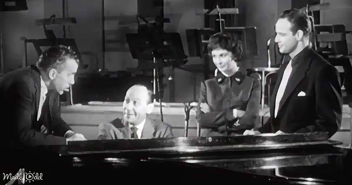 A man in a suit sits at a piano, smiling, with three others around him, in a dimly lit room.