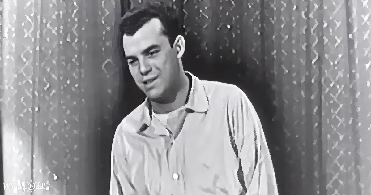 A man in a light-colored shirt stands in front of patterned curtains, looking to the side with a slight smile.