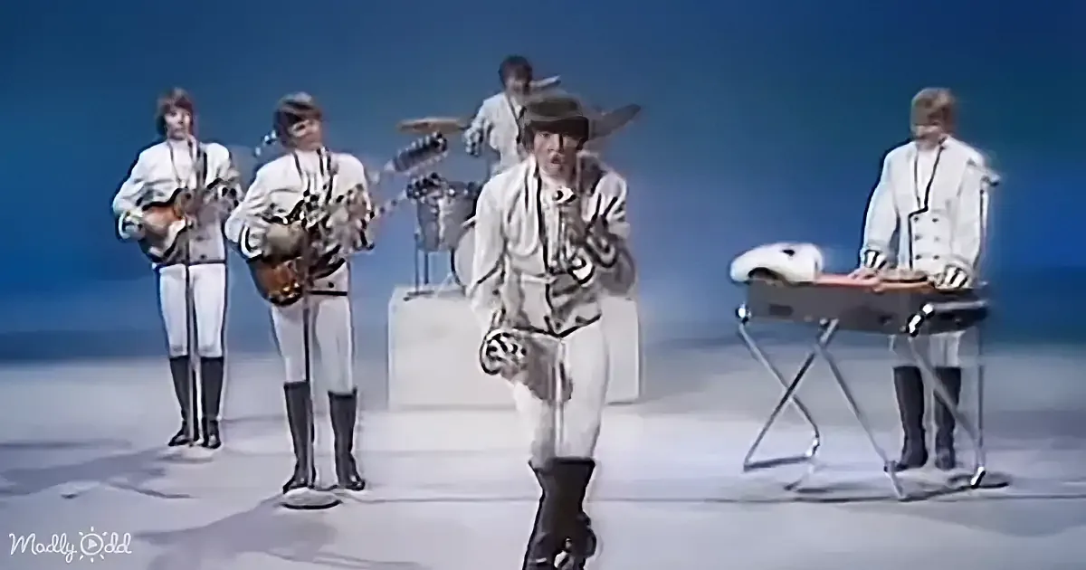 A band in white outfits performs on stage with a blue background, playing instruments.