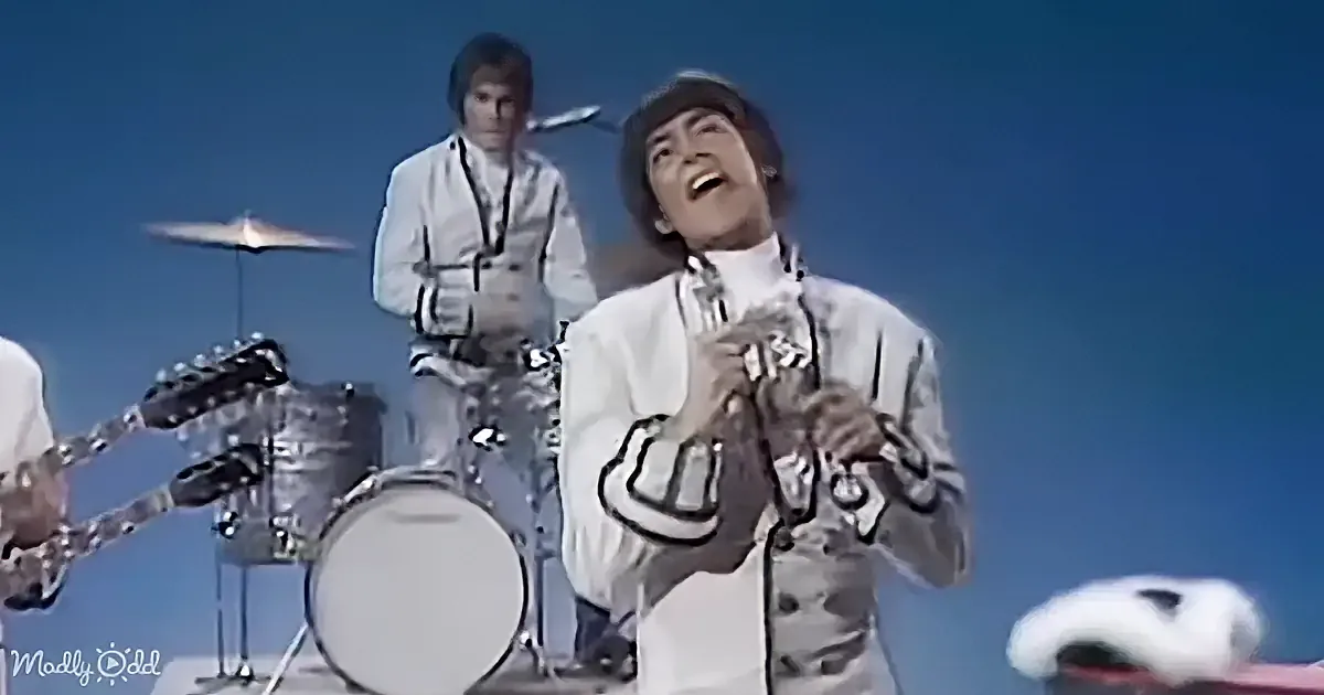 A person in a white outfit and hat is playing a trumpet, smiling, with a drummer and guitarist in the background.