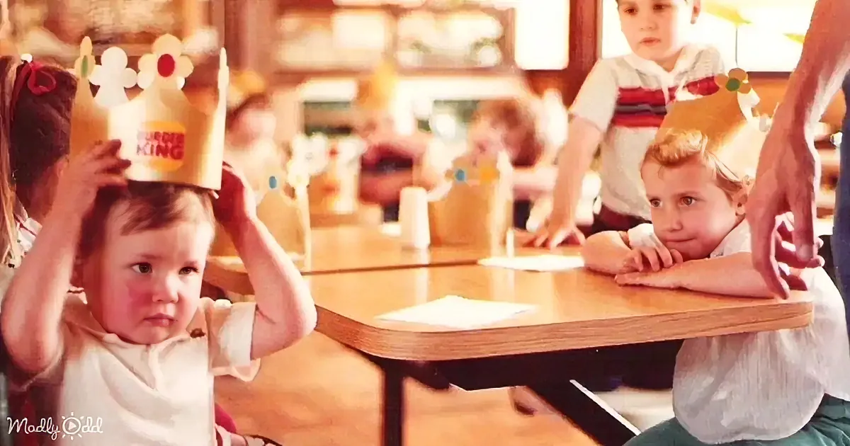 A baby adjusts a Burger King crown while sitting at a table with other children in a restaurant.