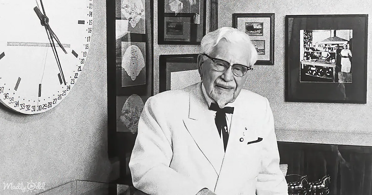 An older man in a white suit and bow tie sits at a desk, smiling, with framed pictures and a clock on the wall behind him.