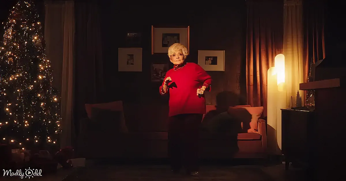 An elderly woman in a red sweater dances in a dimly lit room with a Christmas tree and framed pictures.