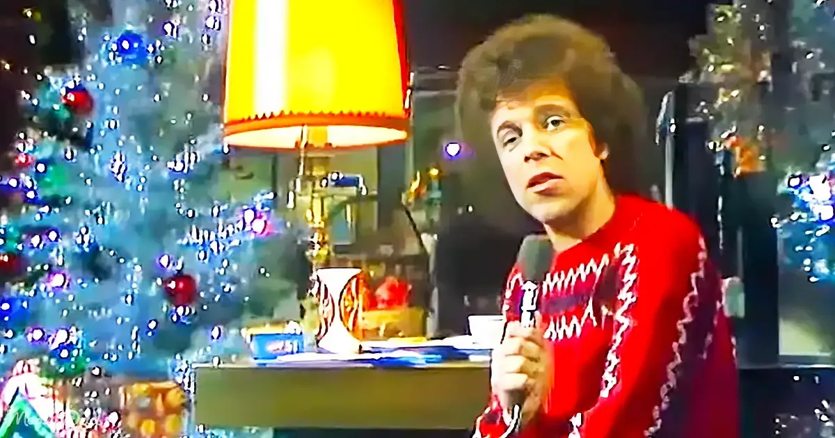 A person in a red sweater holds a microphone, standing in front of a decorated Christmas tree and a lamp.