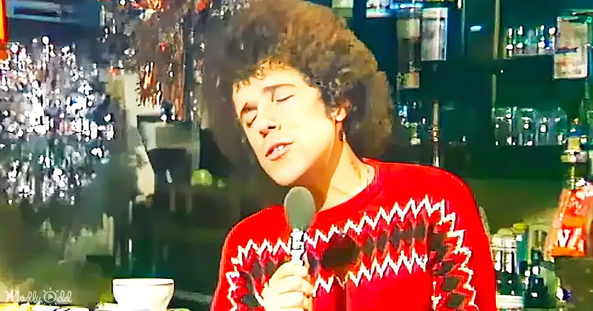 A person in a red sweater holds a microphone, singing with eyes closed, in a dimly lit setting with blurred background.