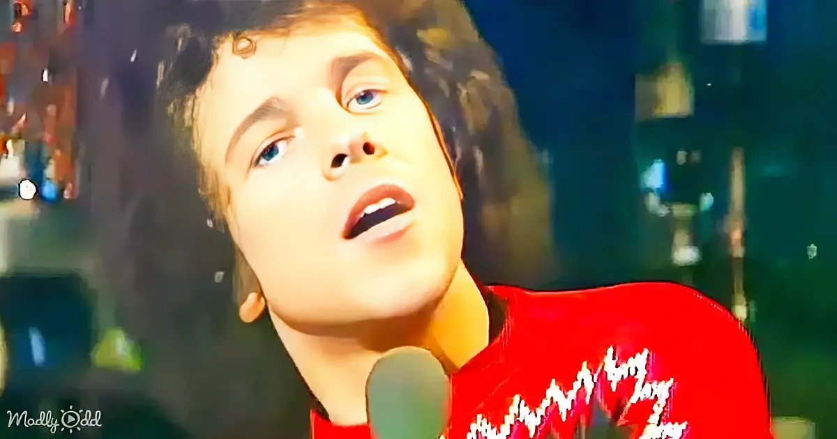 A person in a red sweater sings into a microphone, looking upwards with a blurred background.