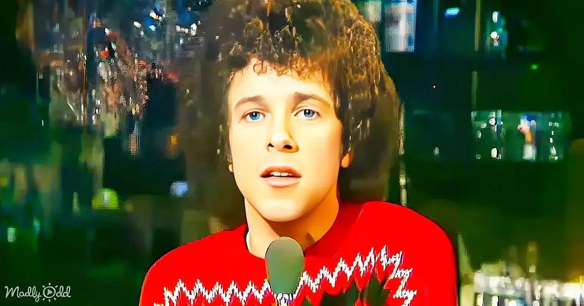 A person in a red sweater with a white pattern stands in front of a microphone, with a blurred background of bottles and...