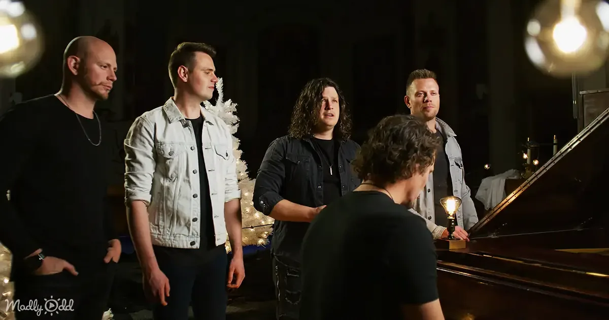 A group of five men stands around a piano in a dimly lit room, with one man playing the piano.