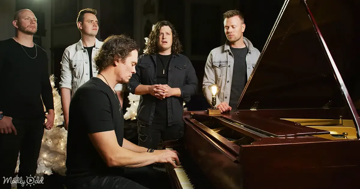 A man plays piano while four others watch, one with clasped hands, in a dimly lit room.