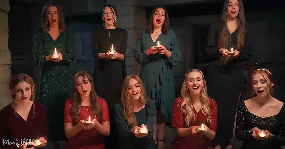 A group of women in elegant dresses hold lit candles, singing together in a dimly lit setting.
