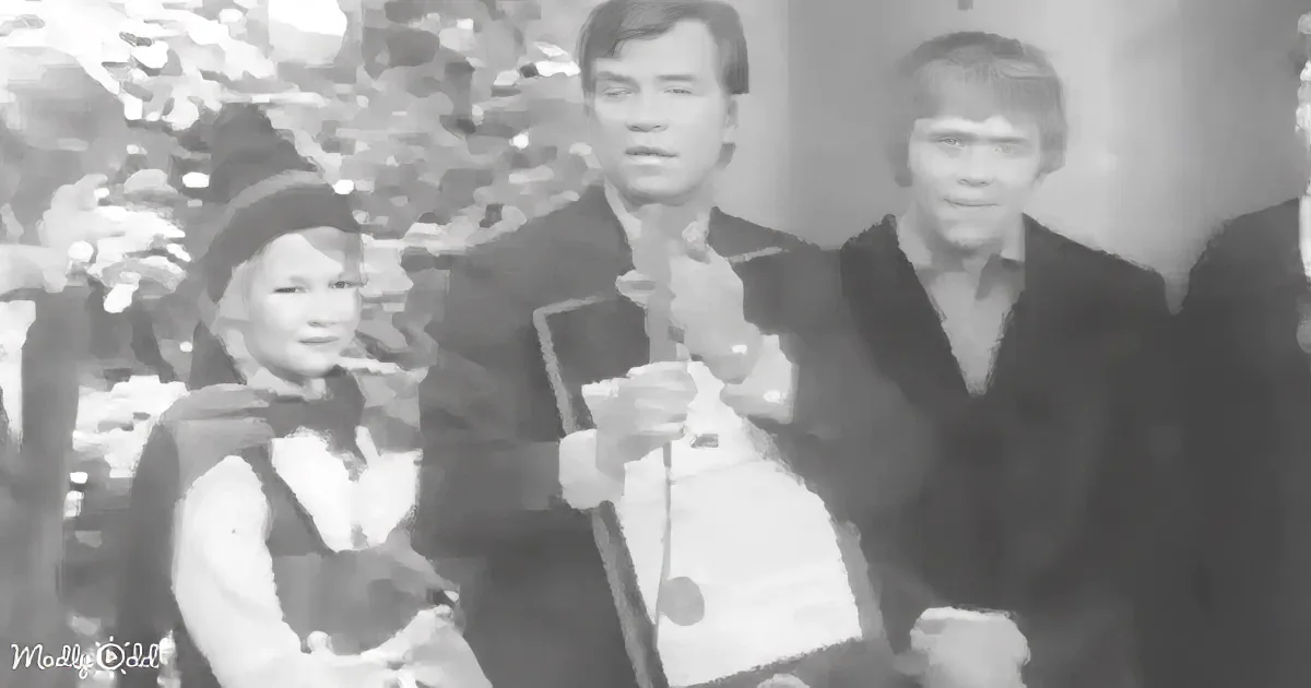 A man in a suit holds a microphone, standing next to a woman in a costume and another man in a dark suit.