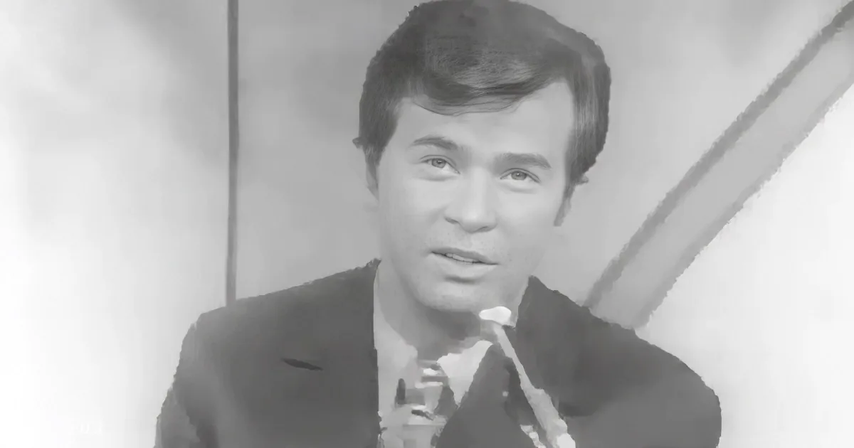A man in a suit and tie speaks into a microphone, appearing to be on a television show or interview.