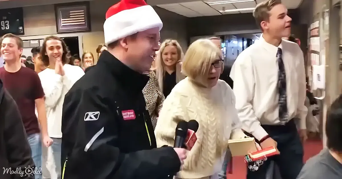 An elderly woman in a white sweater adjusts her glasses while being interviewed by a reporter in a gymnasium.