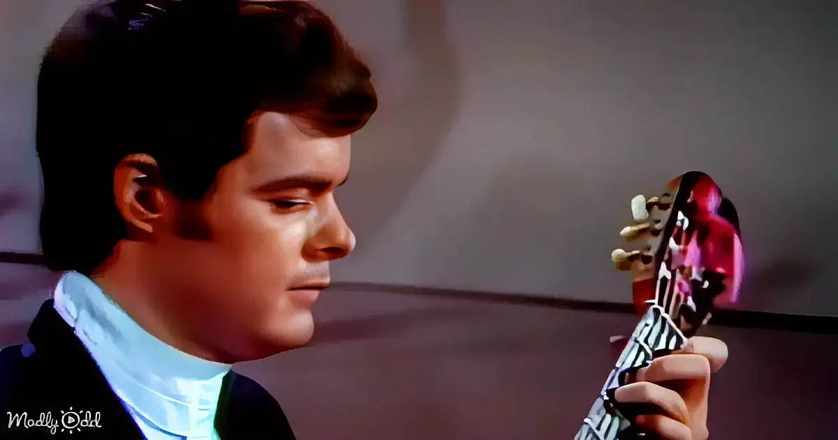 A man in a suit holds a guitar, looking down with a focused expression.