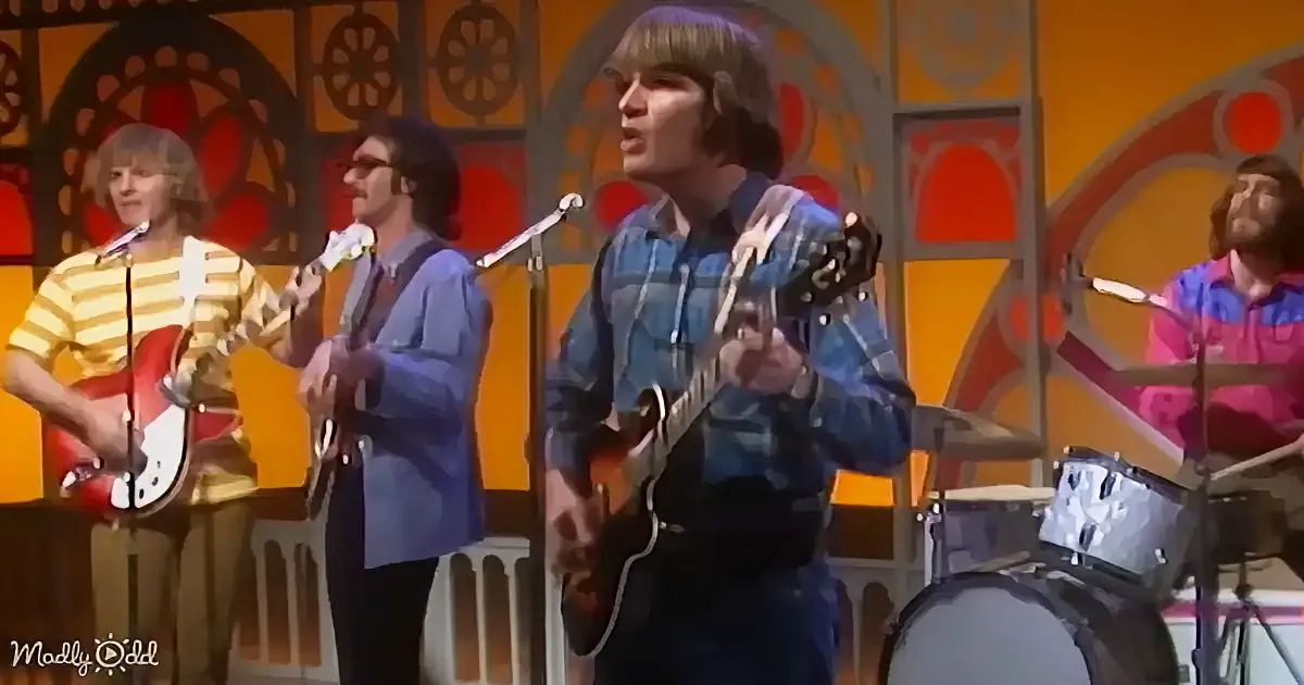 A man in a blue plaid shirt plays guitar while singing on stage with a band.
