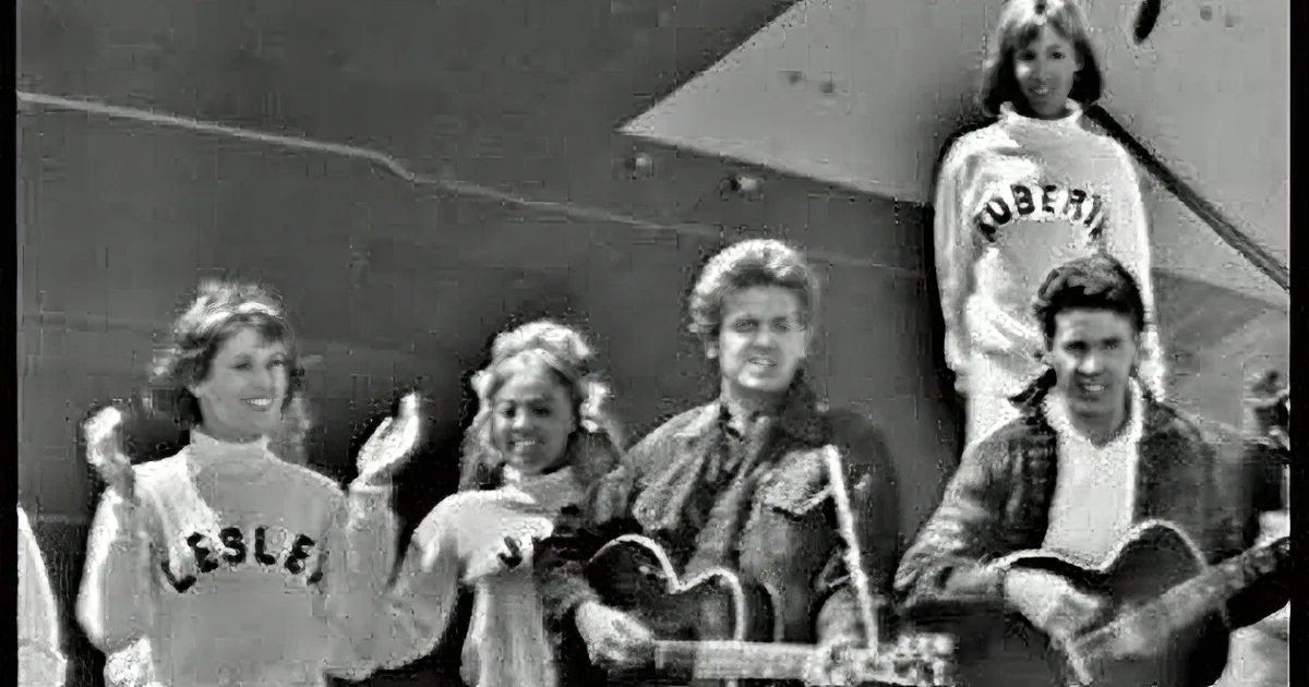 A man in a black jacket and white shirt plays an acoustic guitar, smiling, with another person behind him.