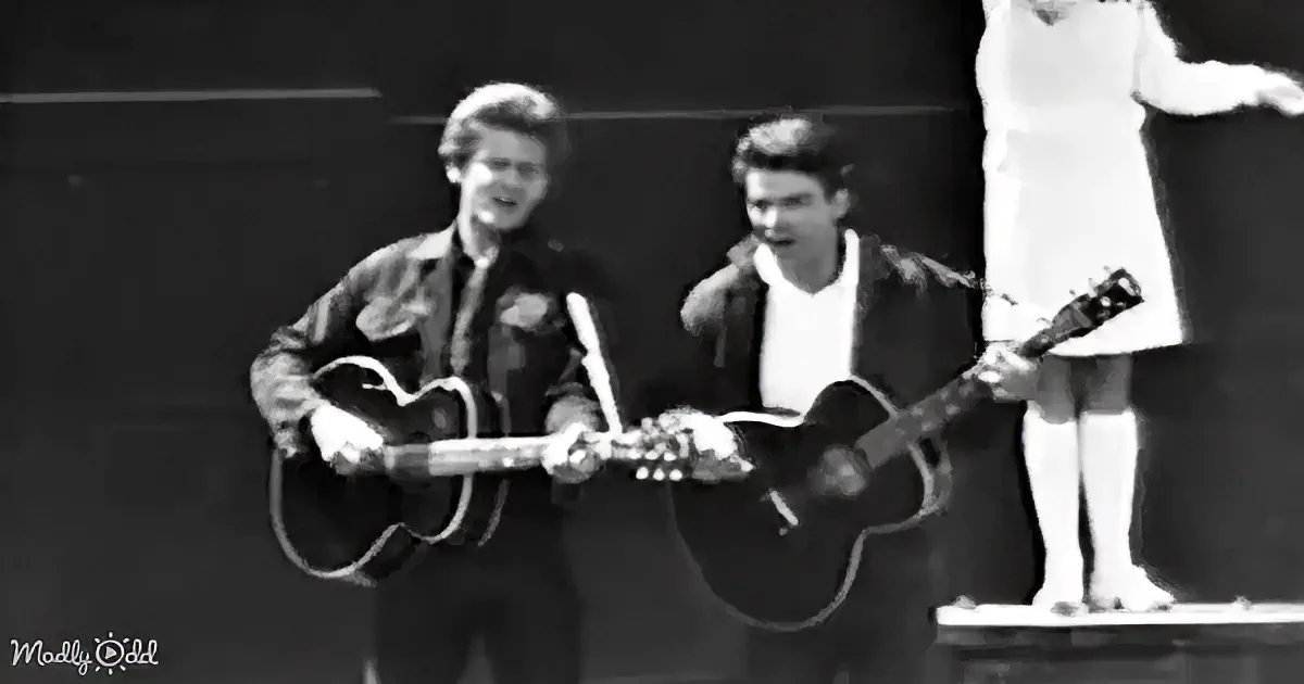 Two men play guitars on stage, one in a jacket, the other in a white shirt, with a woman in a white dress standing behind...