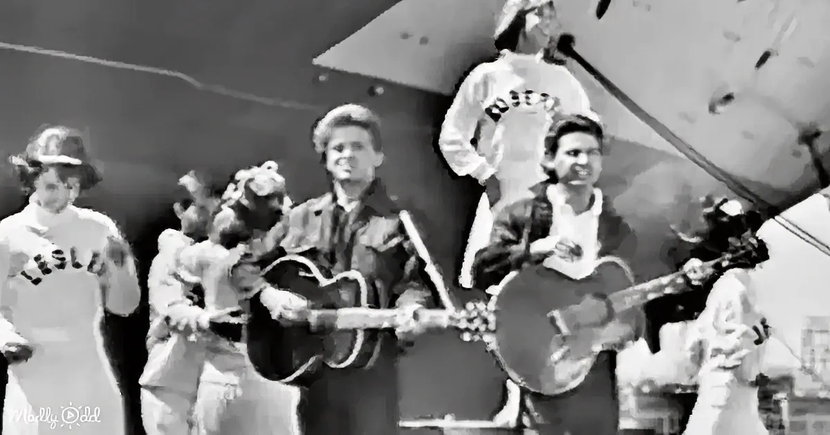 Two men play guitars on stage, surrounded by people in casual attire, with a large aircraft in the background.
