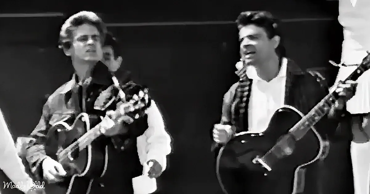 Two musicians play guitars on stage, one singing into a microphone, both focused on their performance.