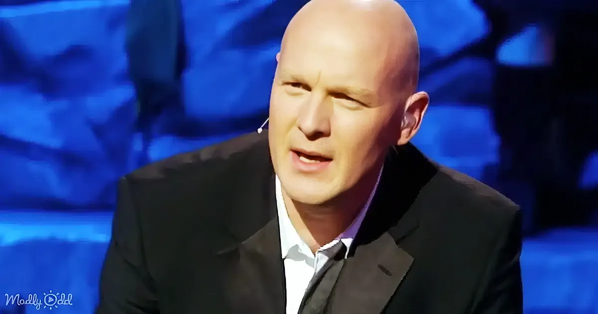 A bald man in a black suit and white shirt appears to be speaking or reacting, with a microphone visible on his ear.