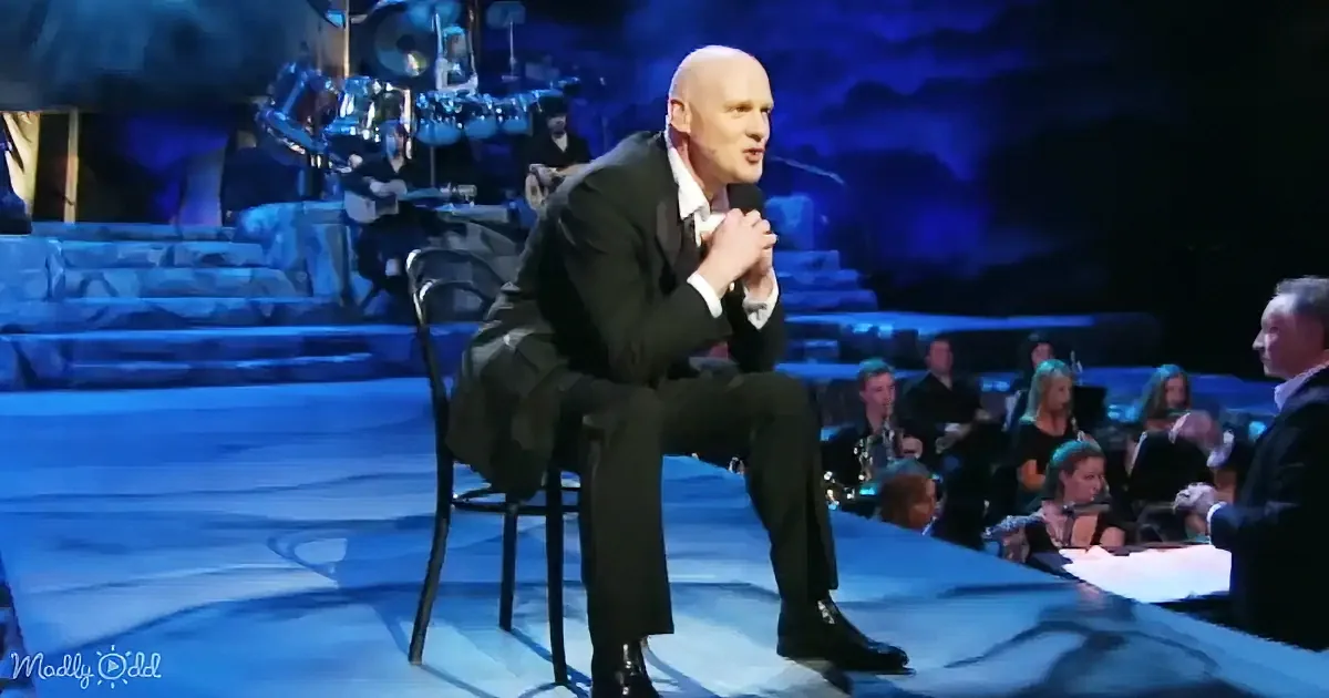 A man in a suit sits on a chair on stage, holding a microphone, with a band and audience in the background.