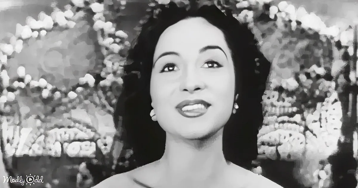 A woman with dark hair and makeup, wearing a dress, smiles against a floral backdrop.