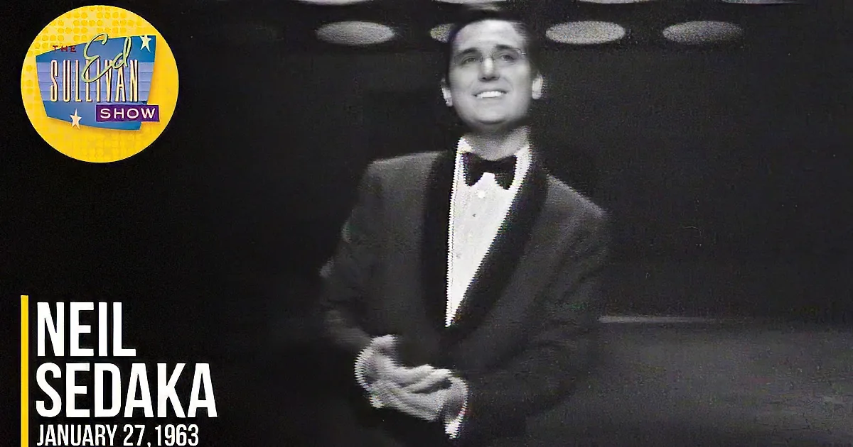 A man in a tuxedo smiles brightly in a dimly lit room with circular ceiling lights.