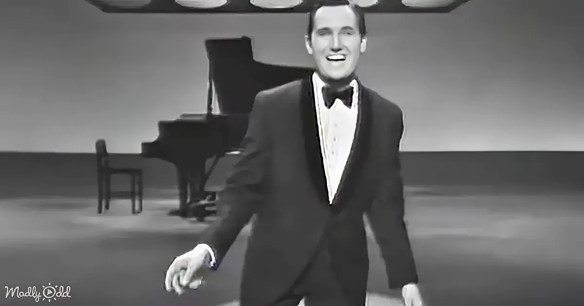 A man in a tuxedo smiles on stage, with a piano in the background.