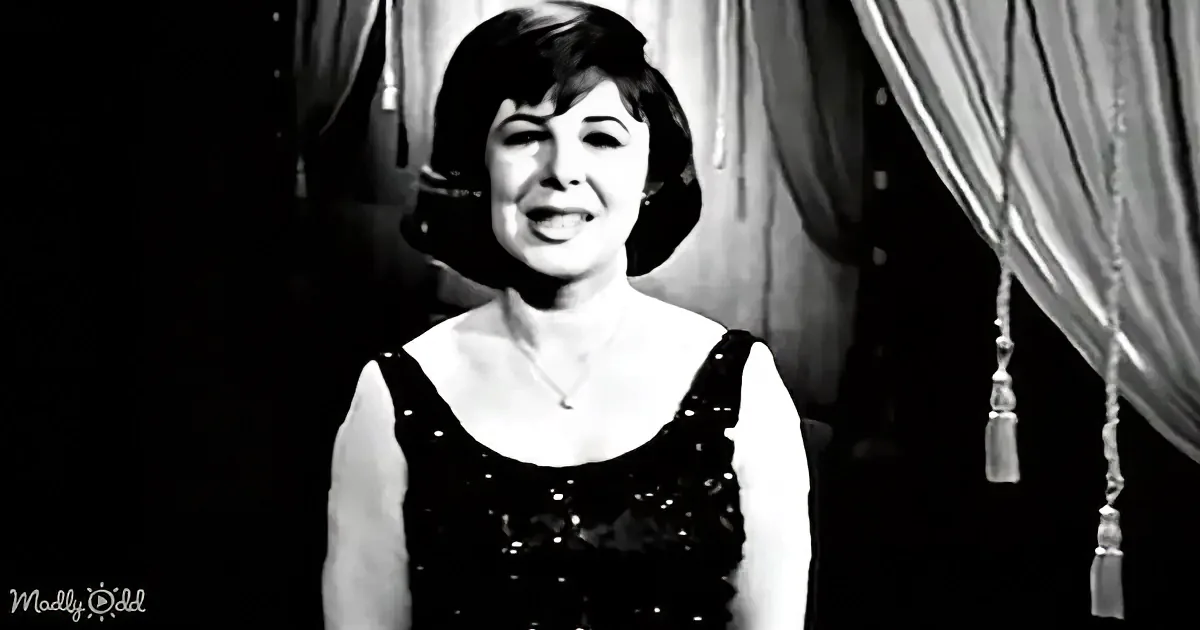 A smiling woman in a black dress stands in front of curtains, wearing a necklace.