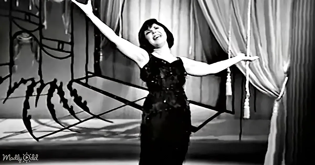 A woman in a black dress smiles, arms outstretched, on a stage with a decorative backdrop.