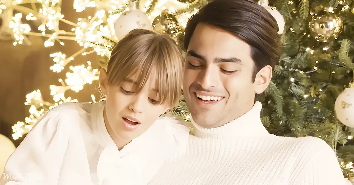 A couple in white sweaters smiles near a Christmas tree adorned with lights and ornaments.