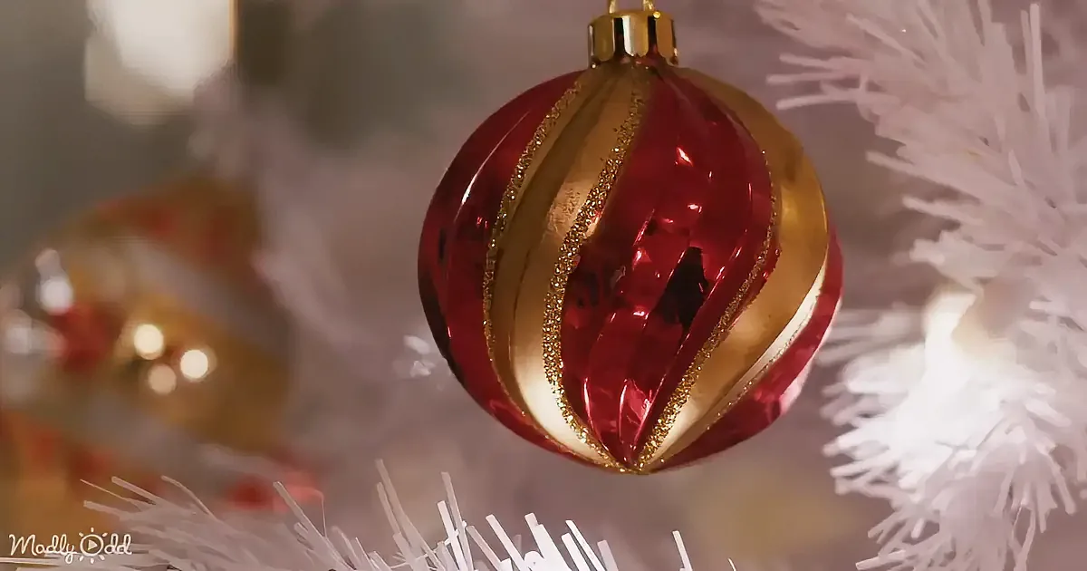 A red and gold Christmas ornament with glittery stripes hangs on a white tree branch.
