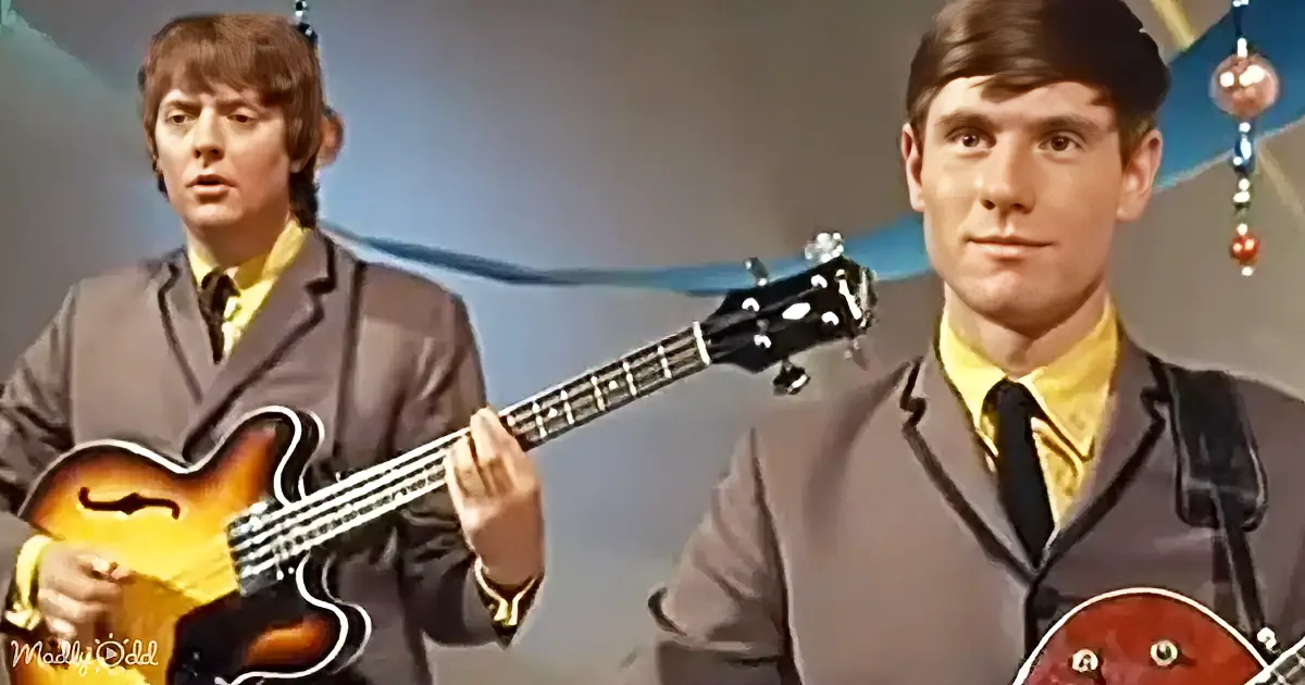 A man in a suit plays a bass guitar, another man in a suit looks on, both in a studio setting.