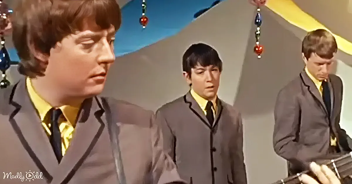 A man in a suit and yellow shirt stands, looking to the side, with two others in similar attire behind him.