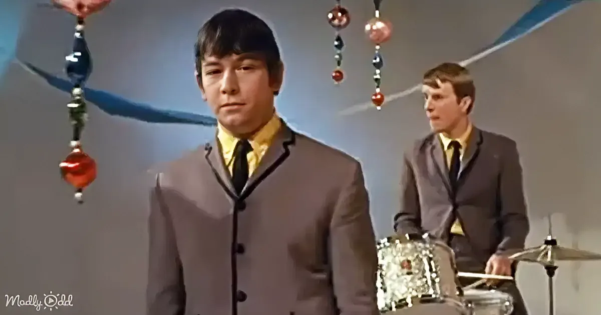 A young man in a gray suit and yellow shirt stands in front of a drummer, with colorful ornaments hanging in the background.