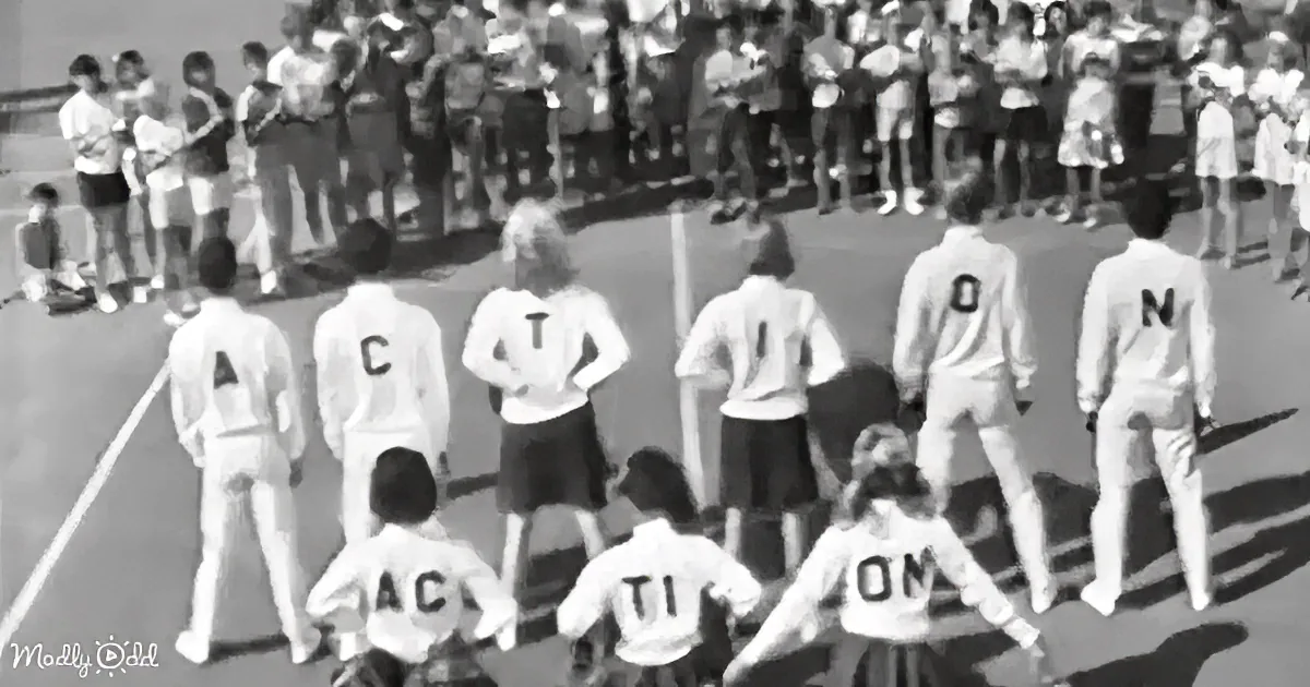 A group of children in white uniforms stands in a line on a field, facing a crowd of spectators.