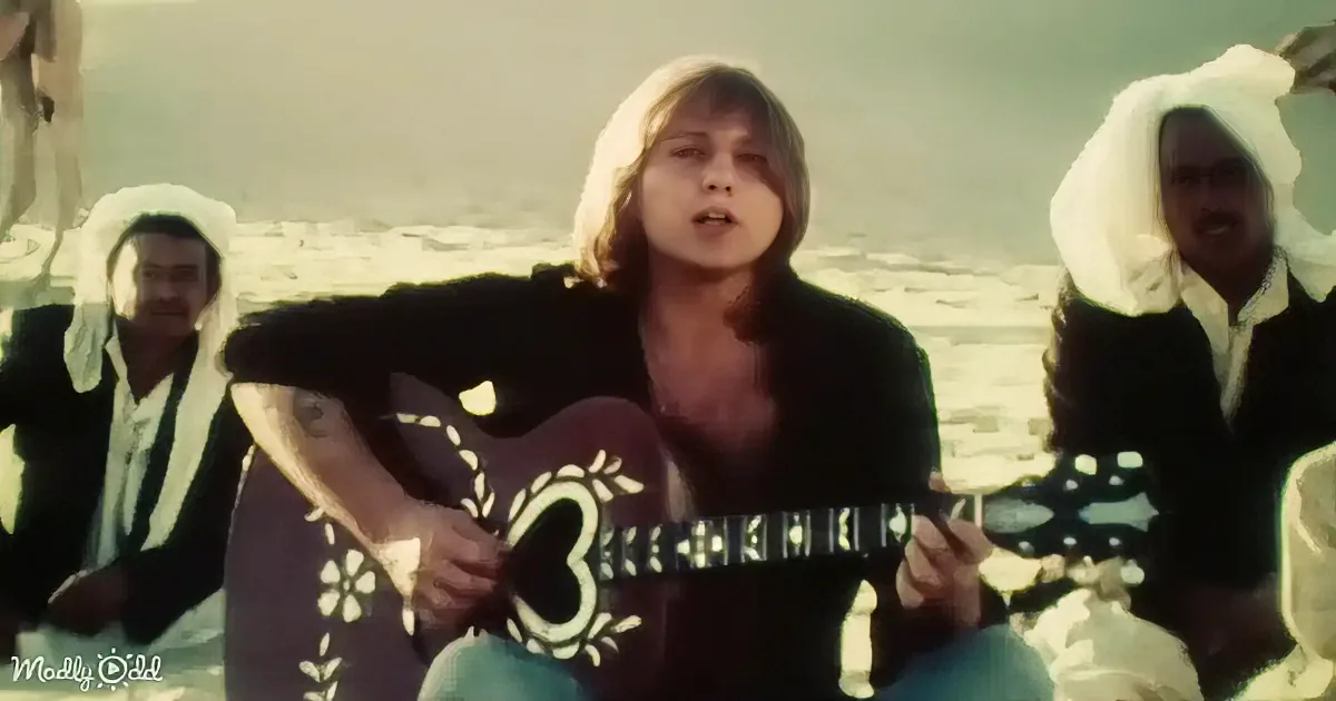 A person with long hair and a black outfit plays a guitar with intricate designs, surrounded by others in white turbans.