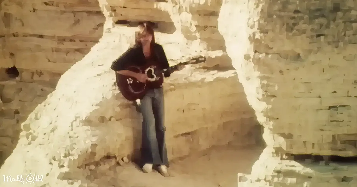 A person sits on a rock, holding a guitar, with a large rock formation behind them.